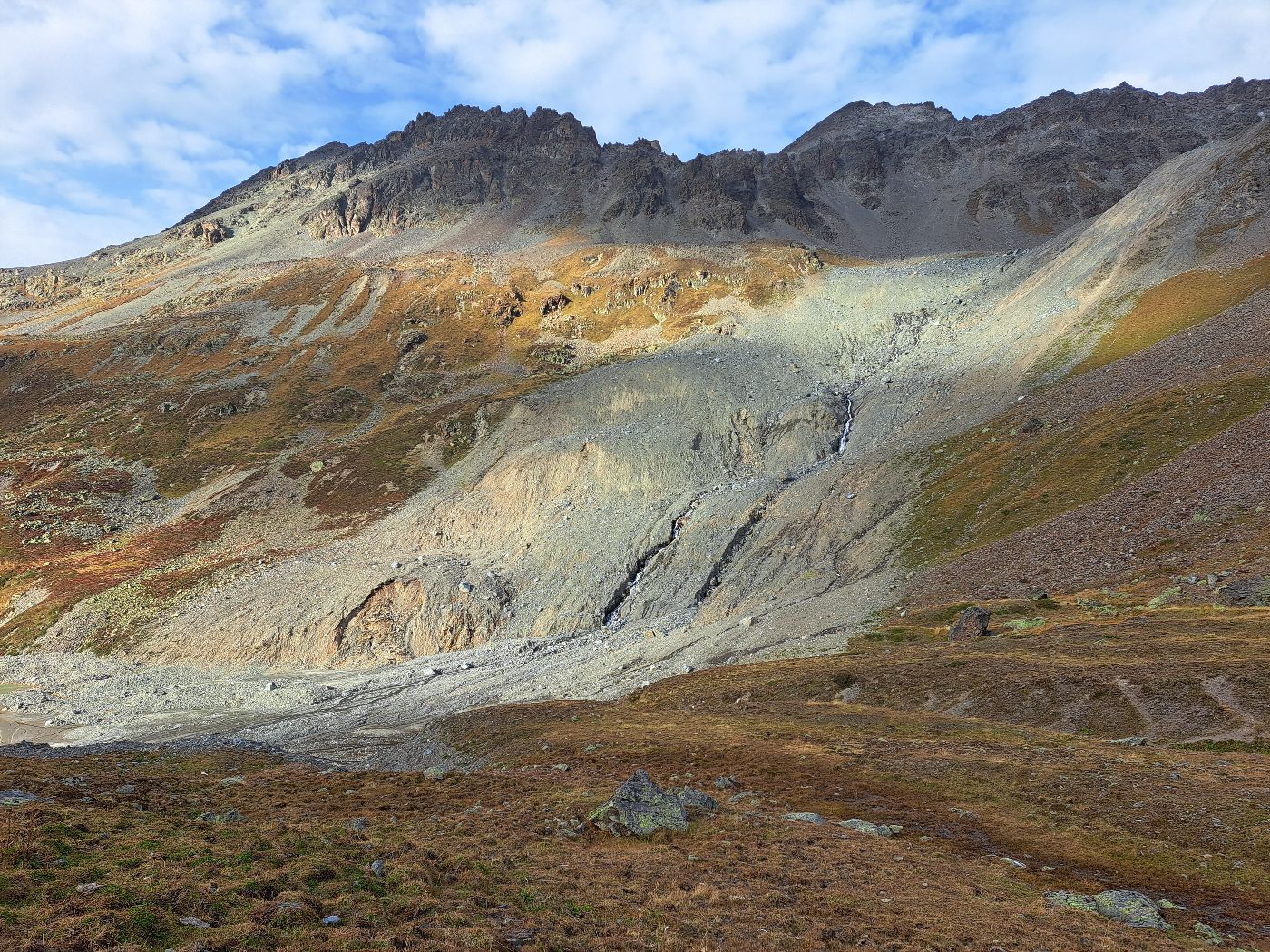 Blick auf karges, alpines Gebiet mit brauner Erde und Bergspitzen, in der Mitte ein grauer Gesteinsstreifen, wo der Bergsturz passierte.