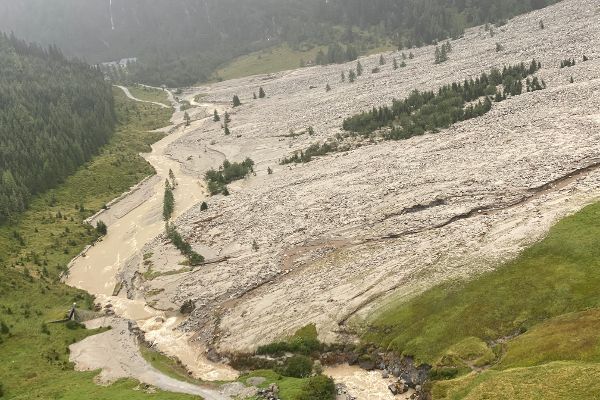 Bergtal mit einem schlammigen Bach, der sich durch das Gelände zieht. Rechts eine große Fläche aus Geröll und Schlamm. Links bewaldeter Hang mit Straße.