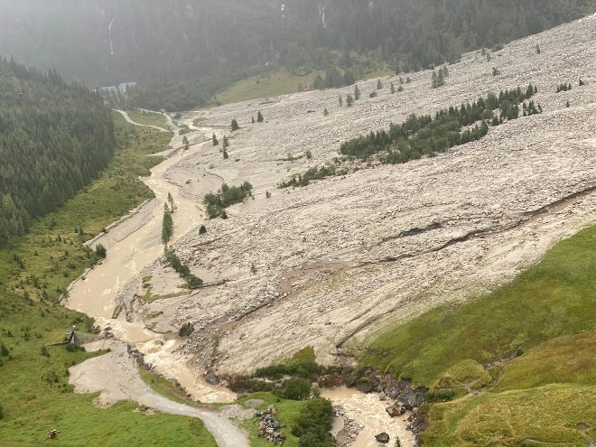 Bergtal mit einem schlammigen Bach, der sich durch das Gelände zieht. Rechts eine große Fläche aus Geröll und Schlamm. Links bewaldeter Hang mit Straße.